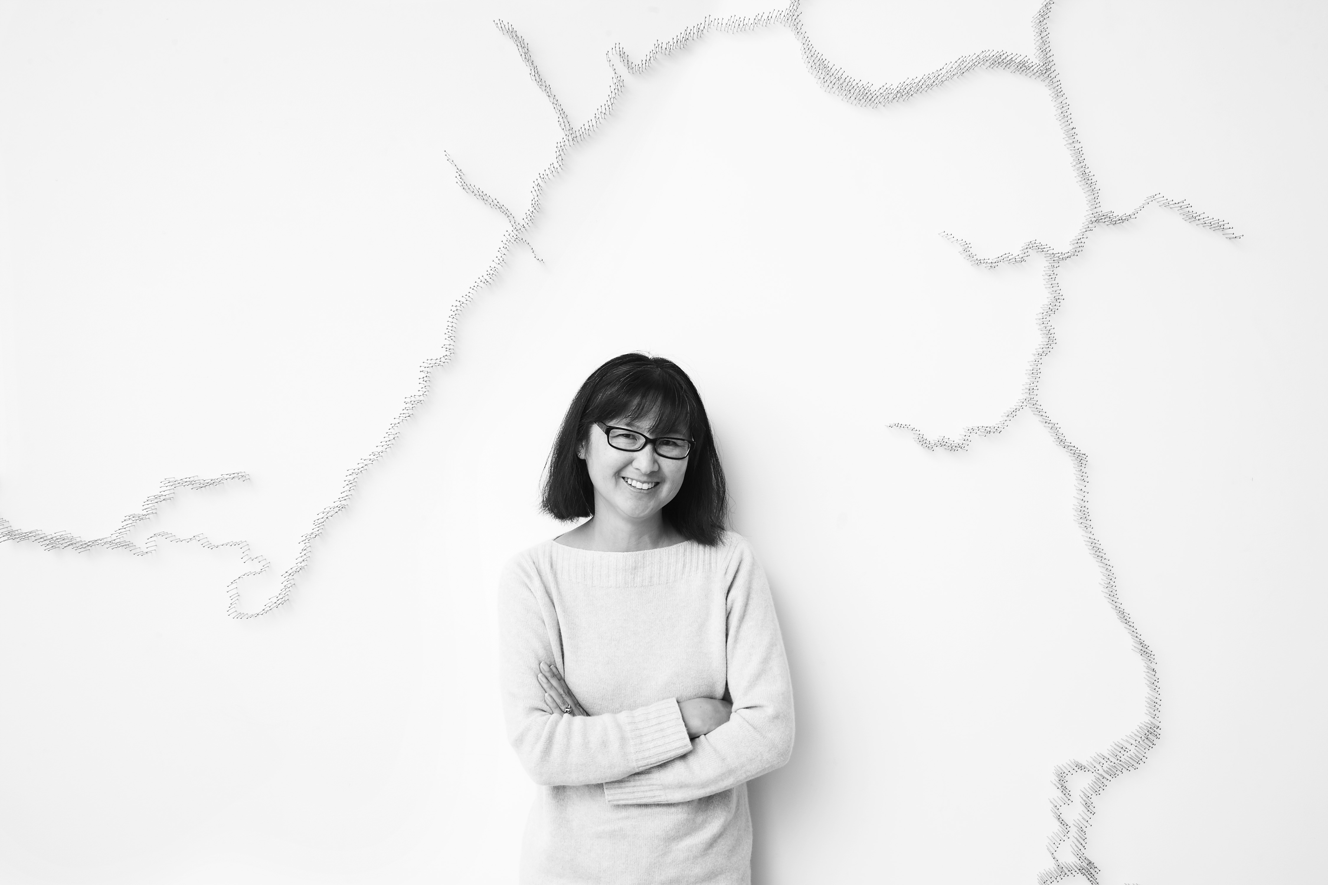 Photo of Maya Lin leaning against a wall that has pins in it in the shape of a river viewed from above