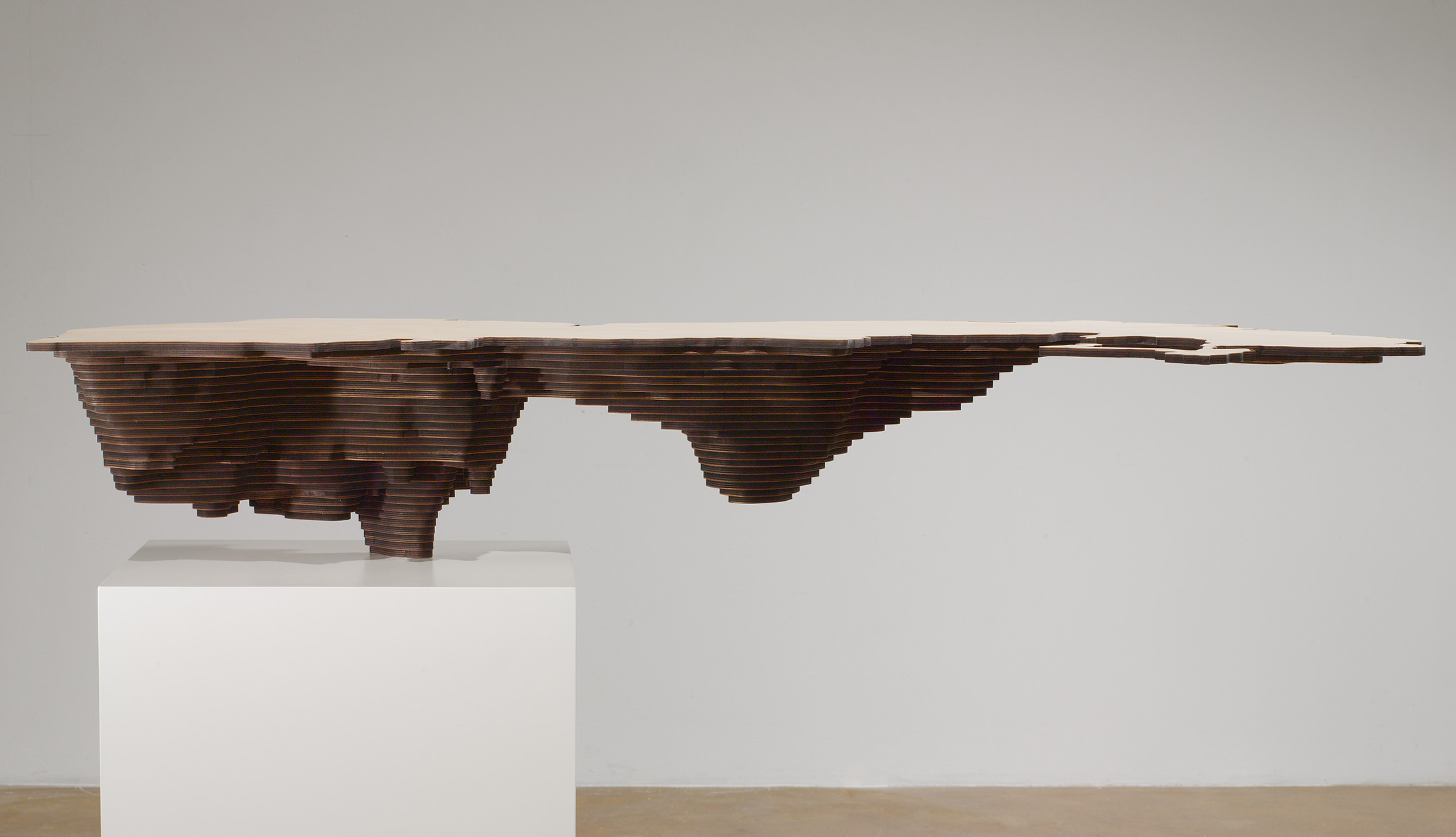 Side view of piece of wood, flat on top with topographical carving of what look like mountains, mounted on a pedestal