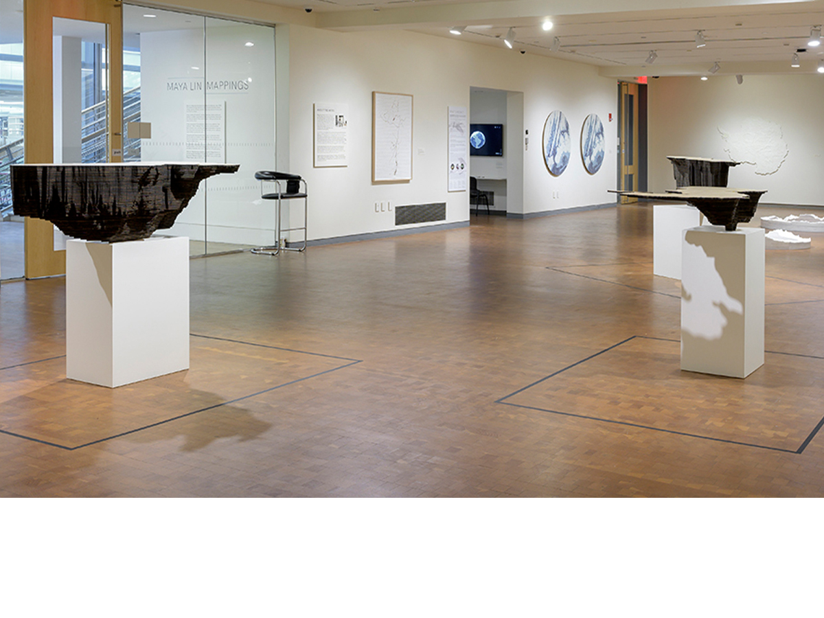 Horizontal image of the Maya Lin Mappigns exhibitio installation with two pieces of pedastal in middle of gallery and artwork on the walls