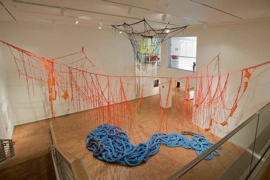 Red ropes hanging from a gallery ceiling with large knotted blue rope laying on the gallery floor