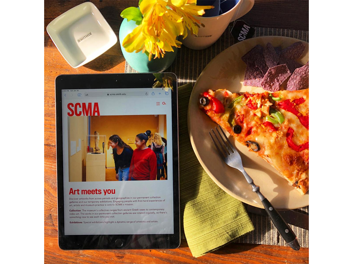 ipad with picture of SCMA website with two students looking at art. ipad on a table with pizza slice and flowers