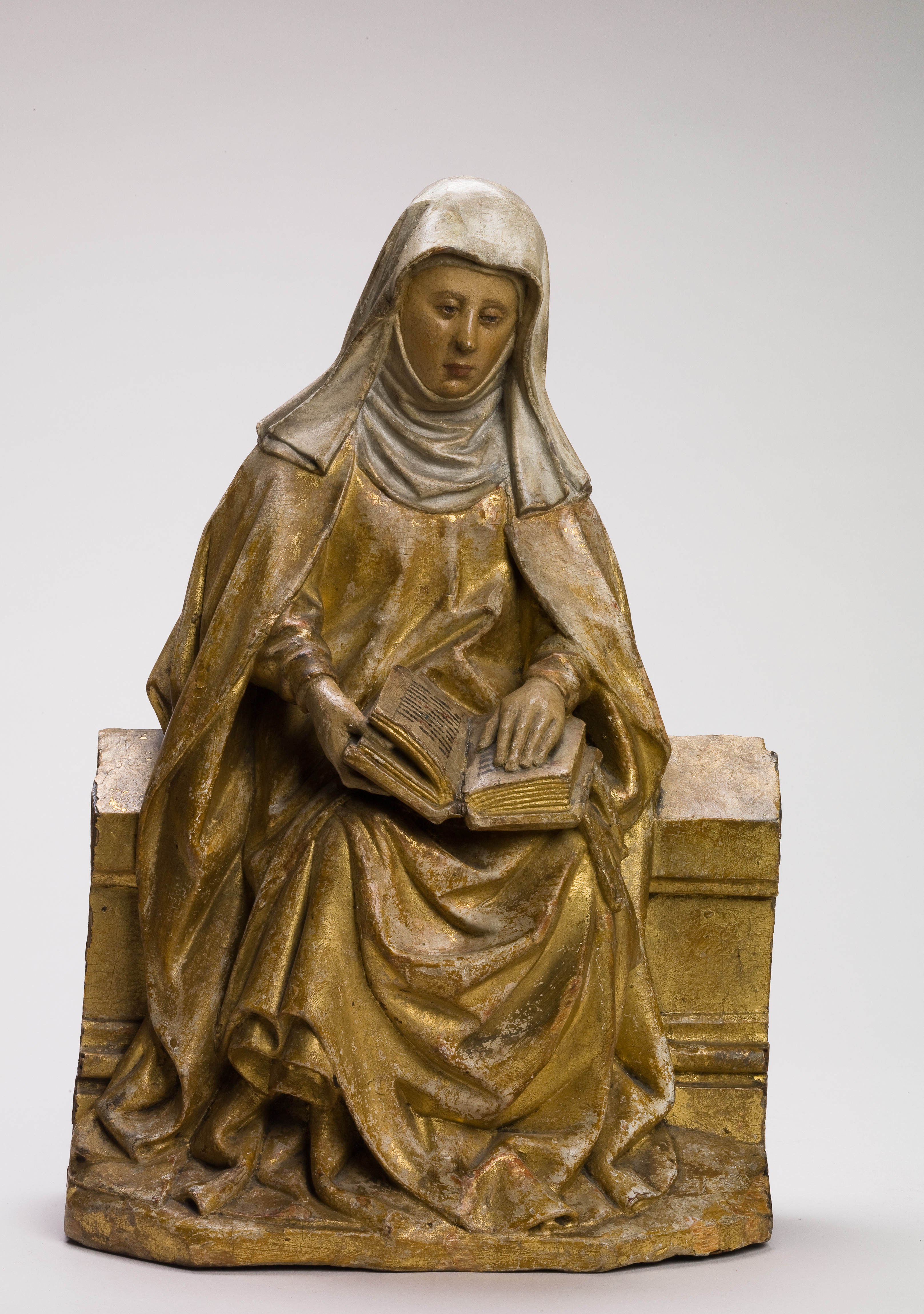 Wooden sculpture of a Saint sitting on a bench reading a book.
