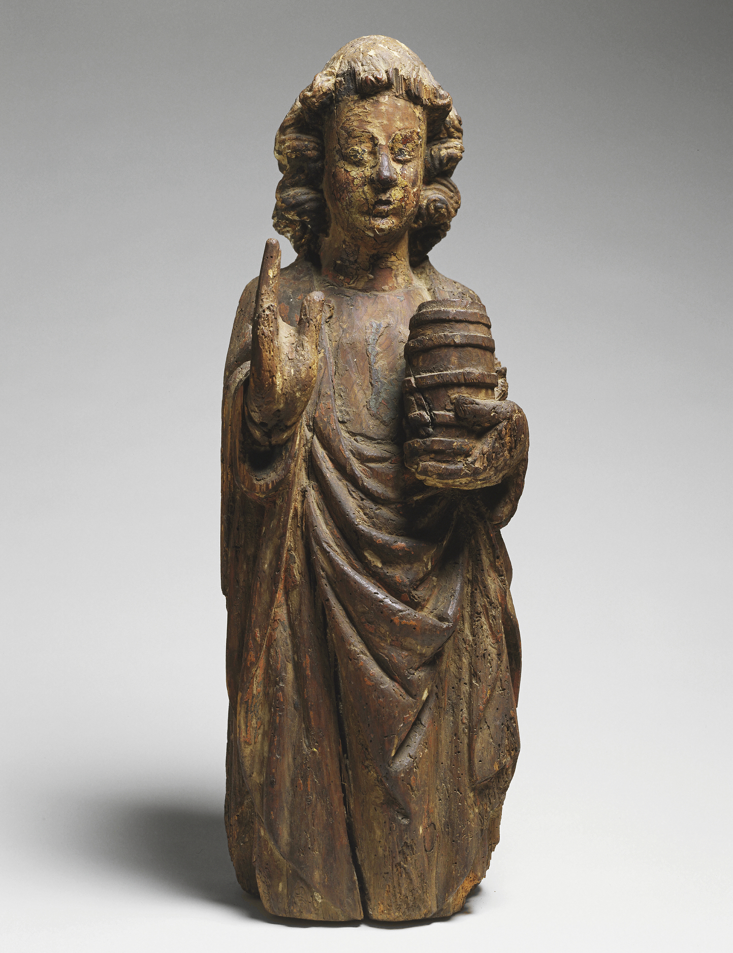 Wooden sculpture of a saint in robes carrying a vessel and holding one hand up.