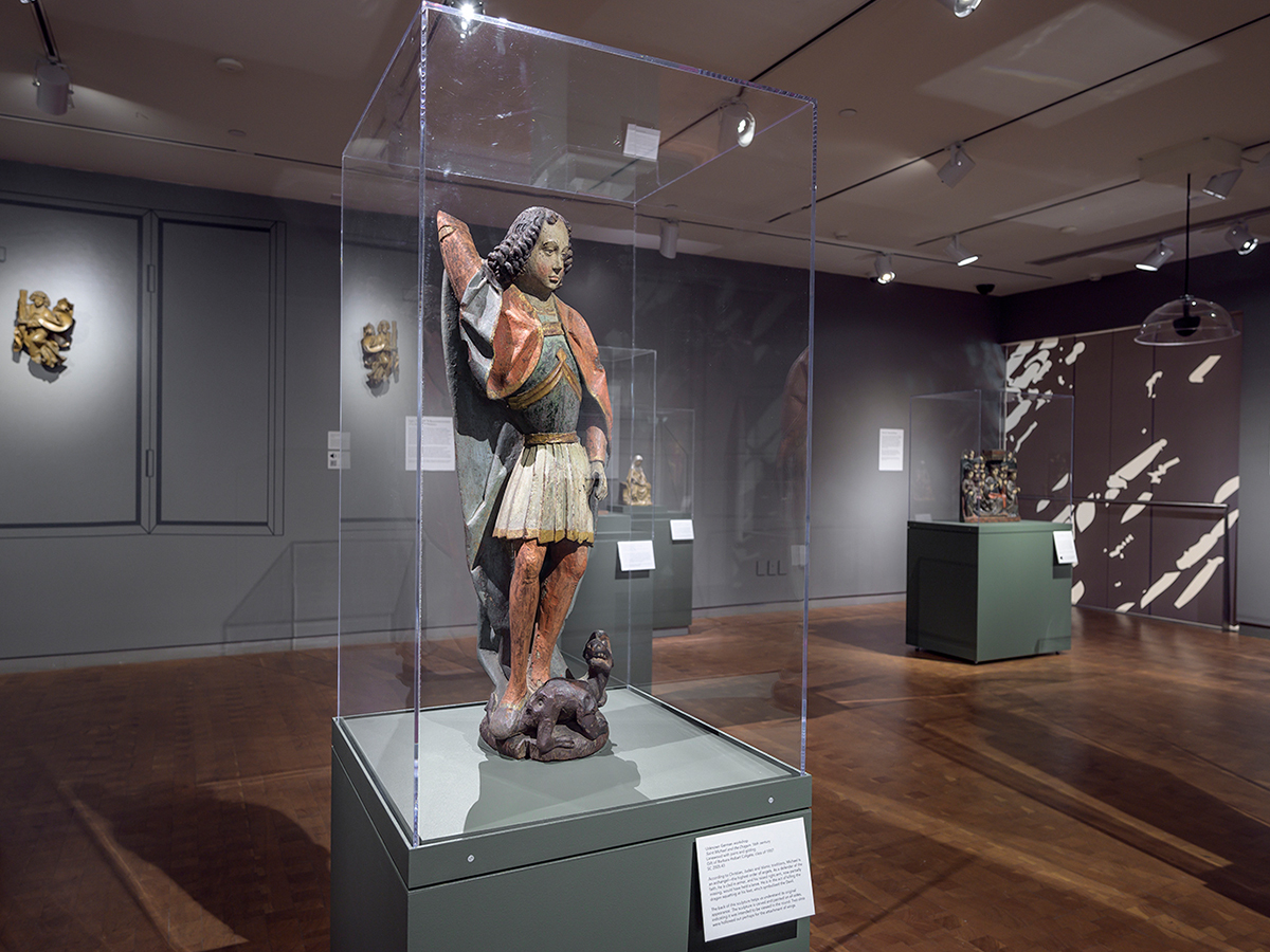 Photo of a gallery with wooden sculpture of a young man reaching for an arrow under a vitrine, with two other sculptures in background on pedastals