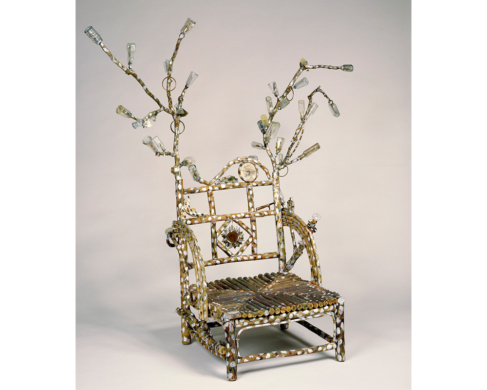 low seated chair made of partly painted branches with curving arms and extended branches at each corner and attached glass, bones, plastic, metal and dried creeper vine