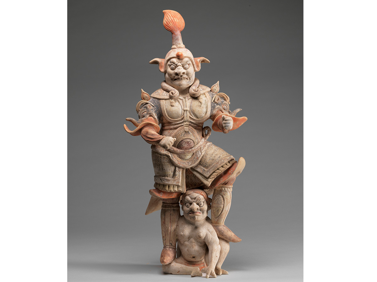 Asian scultpure of a clothed demon standing atop a small unclothed demon made of beige and peach colored stone.