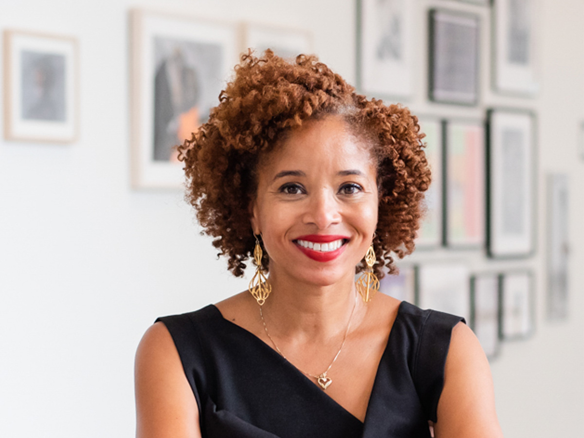 Light brown skinned woman , shoulders up photo, with curly short hair, smiling in black dress sleeveless top with gold earrings and necklace in front of a blurred wall with pictures hanging on it.