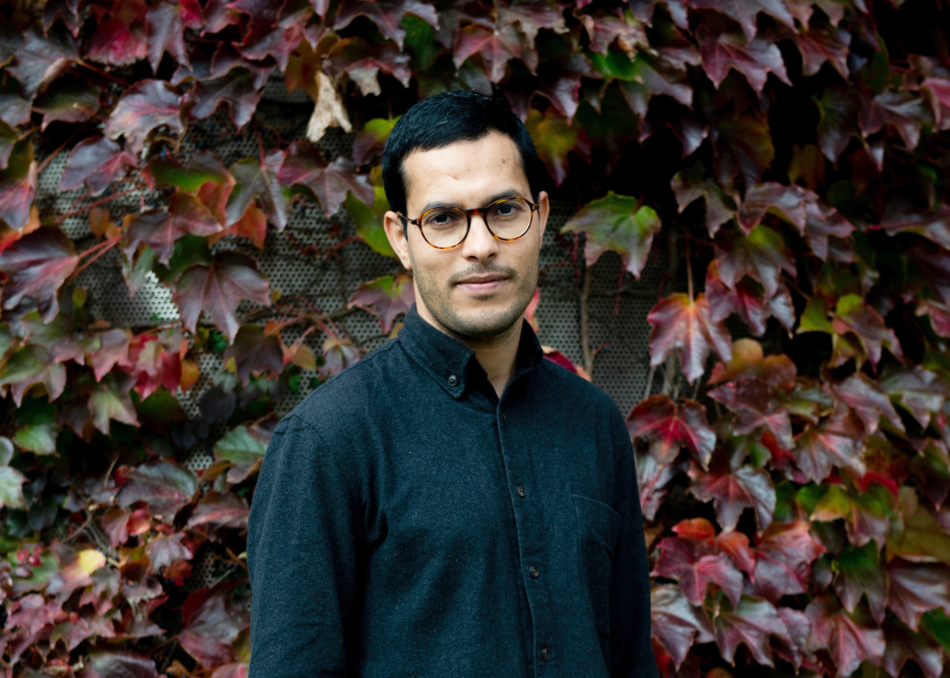 Torso phtoto of a young man with dark hair, glasses in a button down deep teal blue shirt standing in fronta wall of ivy-like vines that have turned colors like red and deep green.