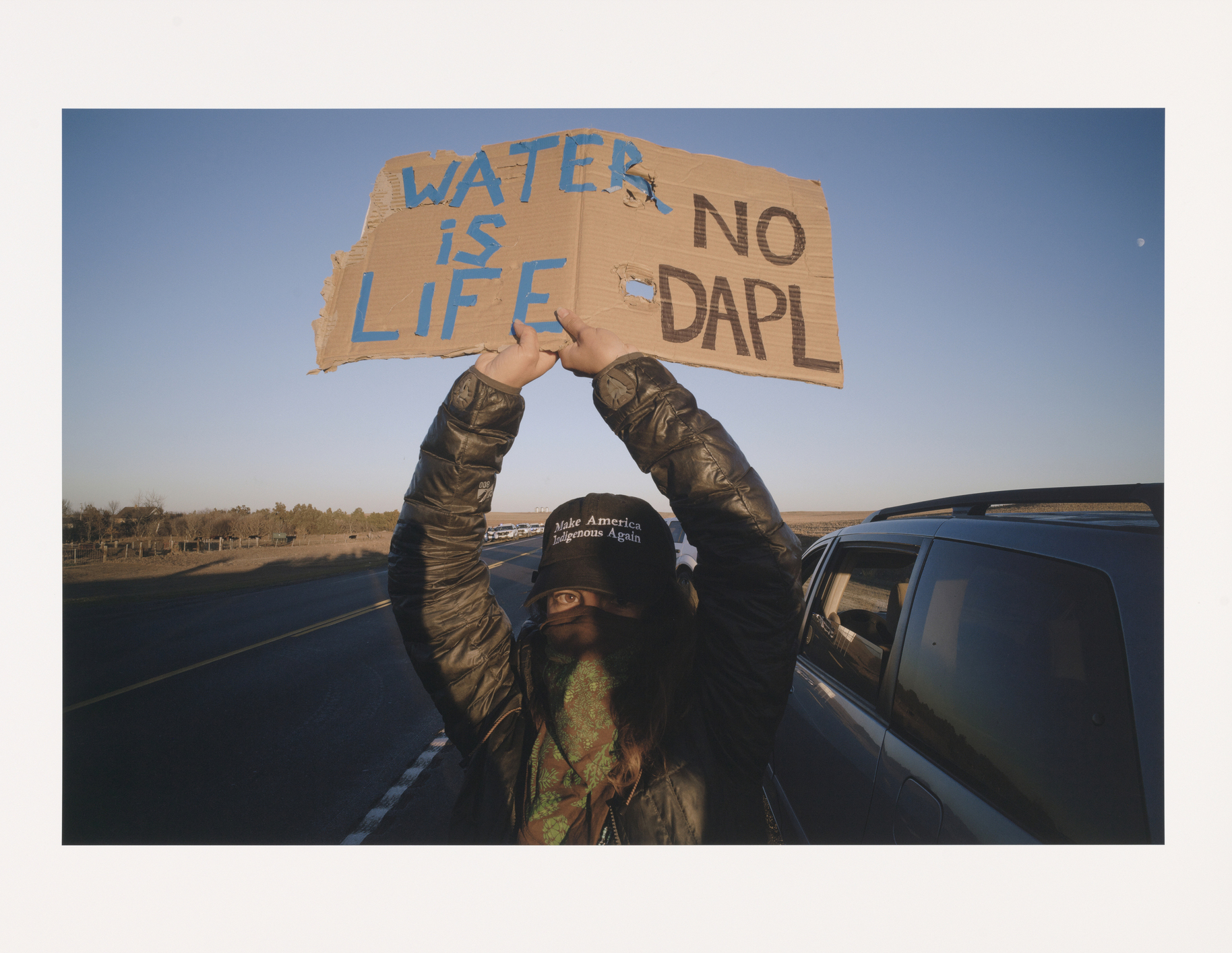 Person standing on the side of a road next to a car, in a leather jacket with scarf over their face with a hat that reads: Make America Indigenous Again, holding cardboard sign over their head that reads "Water is Life," No DAPL