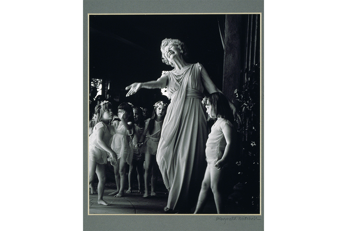 a woman dresssed in an evening gown stands in the middle of a group of children with her arm out streched towards the viewer