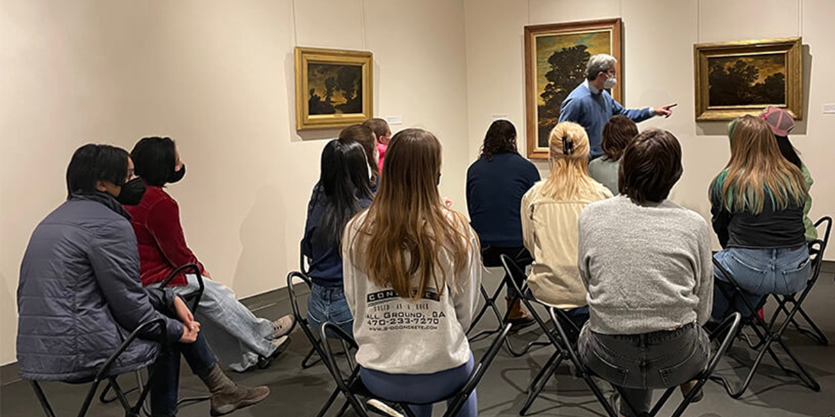 Man in front of a painting in gallery with students in gallery chairs listening and taking notes