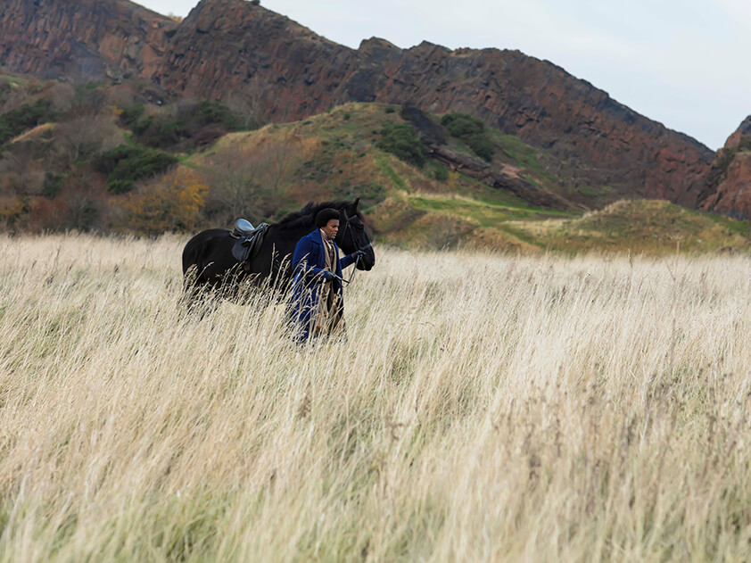 Man dressed in a blue jacket from late 1800 with a horse standing in tall grass with rocky hill in the background