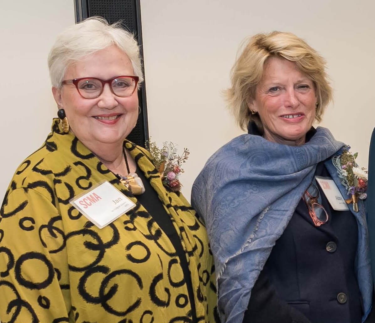 Two older women, one on left has gray hair, red rimmed glasses and a yellow top with black circle design on it. On right is a blond haired woman with blue scarf over a dark blue jacket, both are smiling