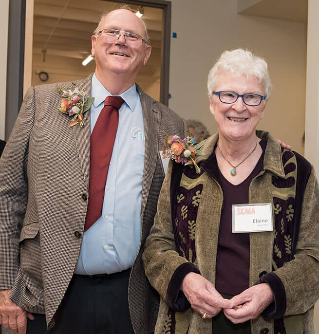 Smiling older gray haired man in a tan suit and red tie stnding next to a smiling older gray/blond haired woman in a vertical striped sweater with her hands touching in front of her