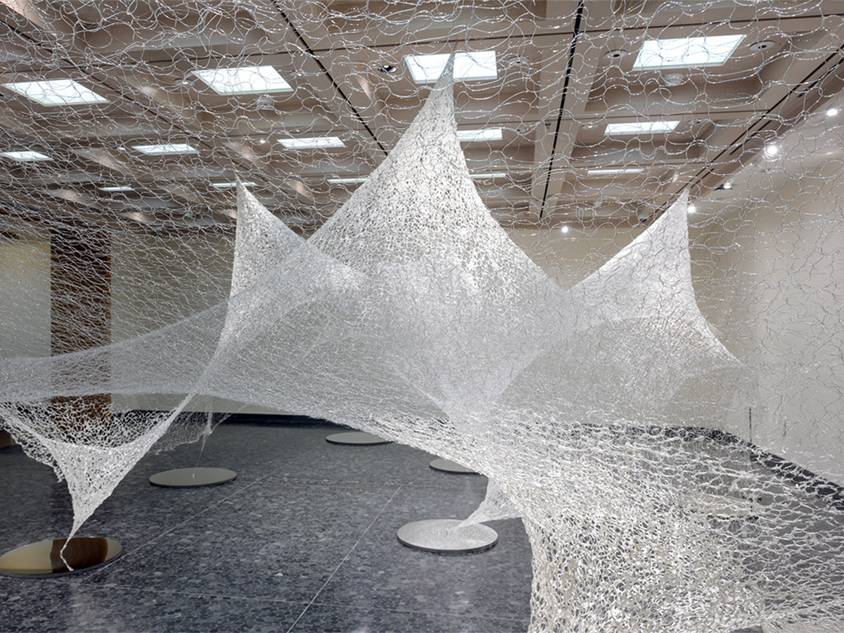 Photo of a shimmering webbed construction hanging in a gallery with recessed ceiling lights and dark stone floor