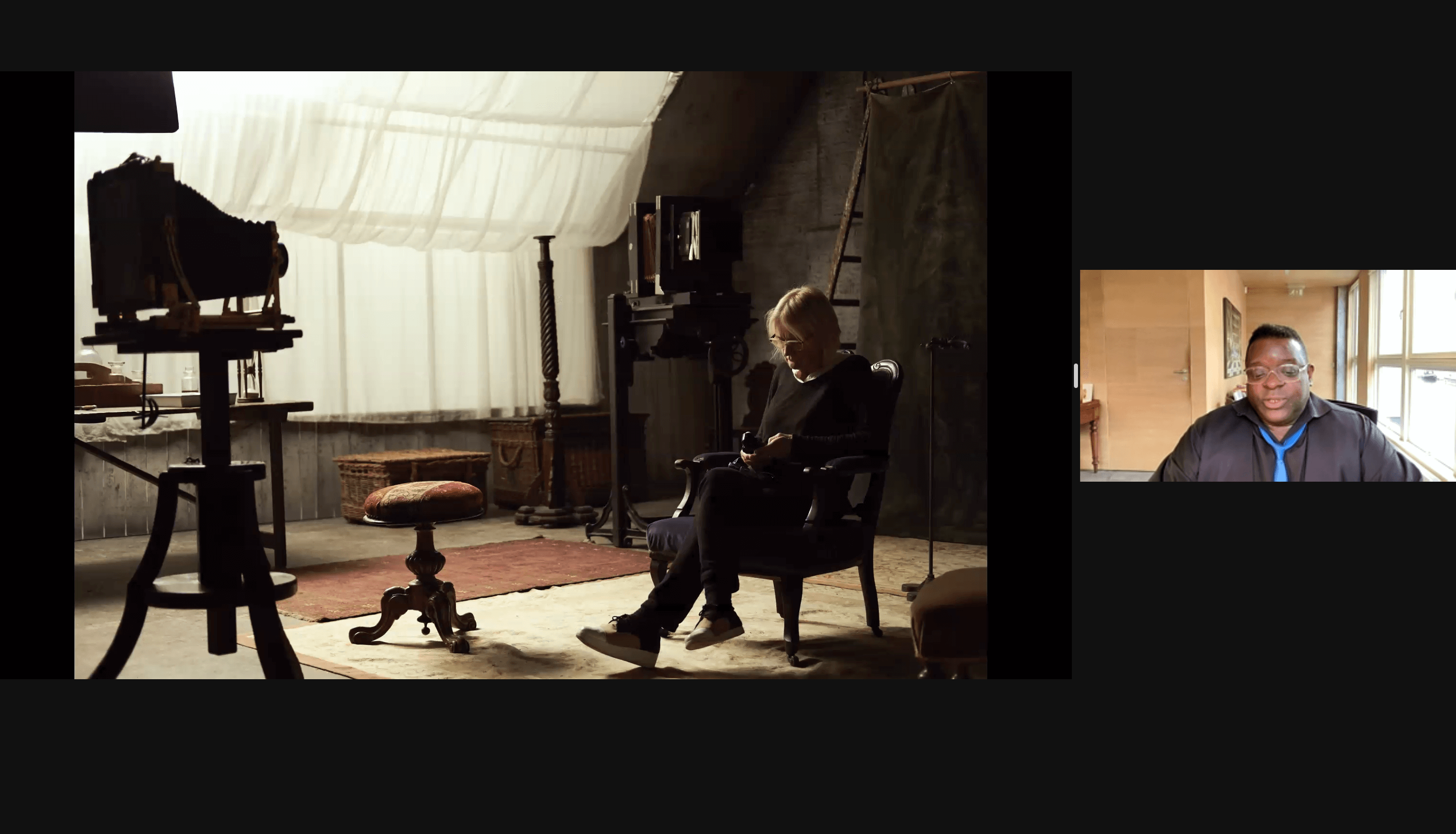 Looks like a screenshot from a Zoom presentation with image on left  of a blond haired person sitting a set with an old photo camera on a wooden tripod, with the Zoom presenter box that includes a man in clear rimmed glasses with short black hair in a black button down shirt and bright blue tie.