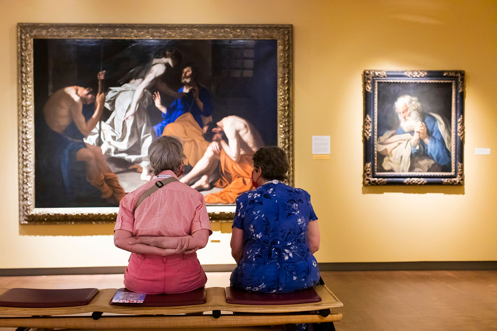 Two older people sitting on a bench in a gallery, with backs to the viewer