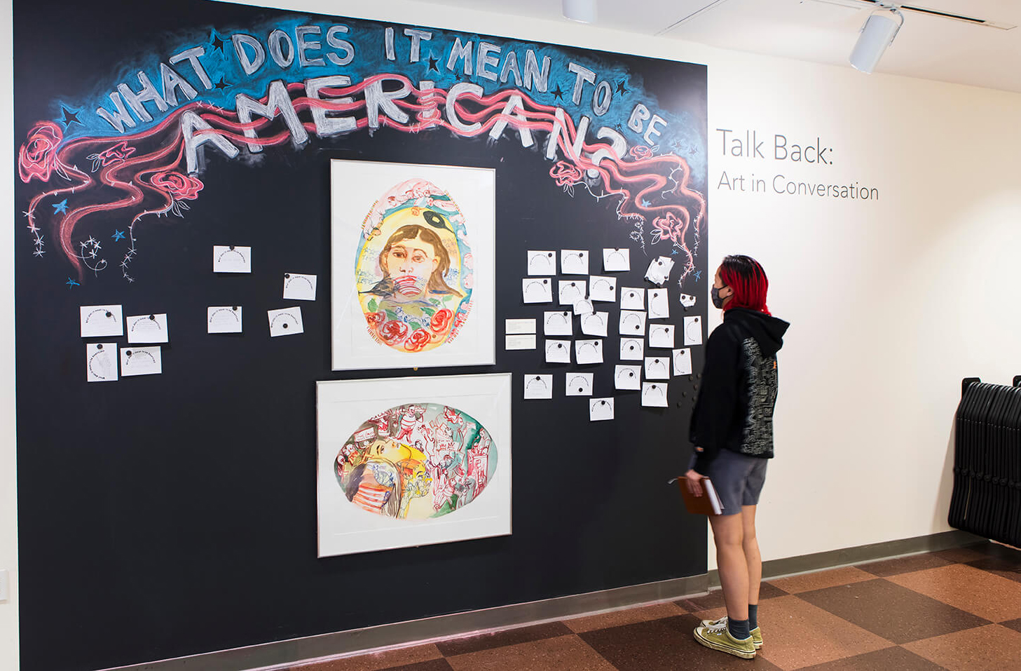 Single student in sweatshirt and shorts standing in front a a wall with the chalked letters that read: What Does it Mean to be American?
