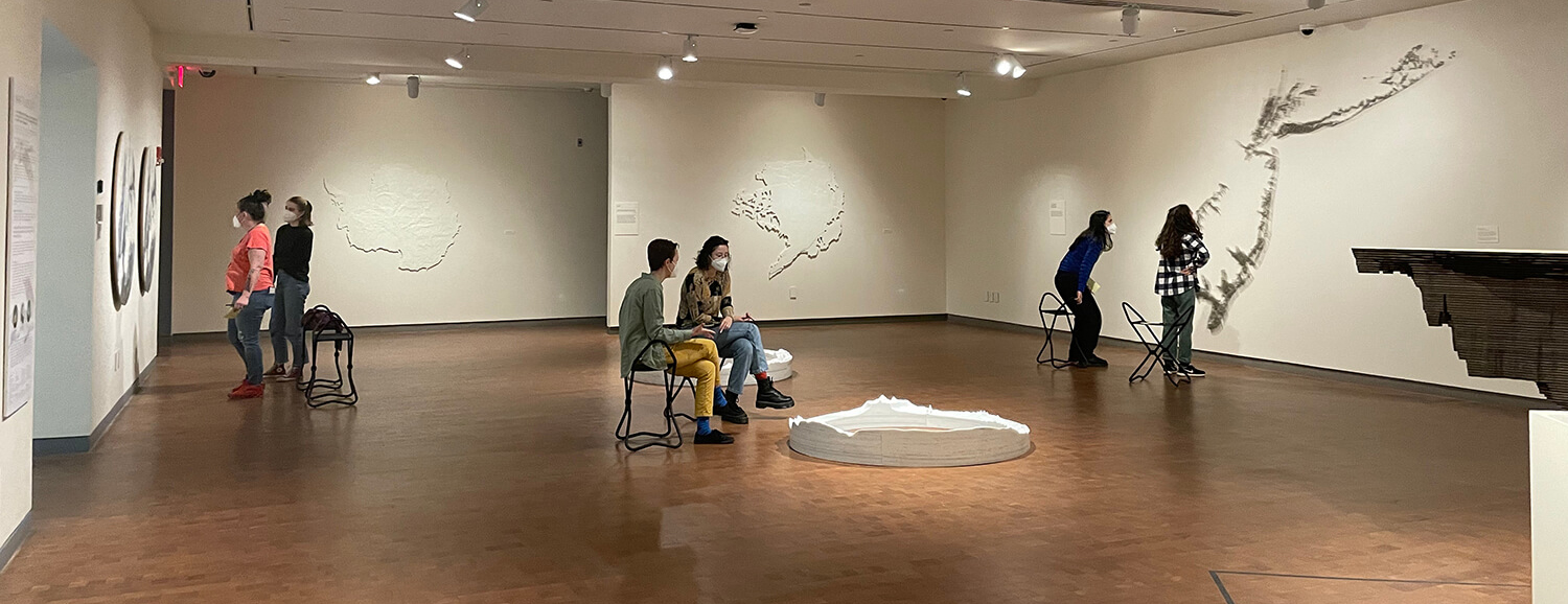 Several student in the distance standing and in chairs in a gallery with white circular sculptures on floor and delicate artworks on the walls