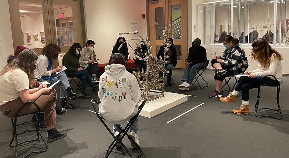 Student sitting in gallery chairs surrounding a sculpture on a pedestal in the middle of the gallery