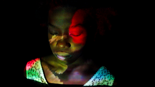 Still from movie with abstract image of a woman in foreground with colorful sleeveless shirt and reflection on her face
