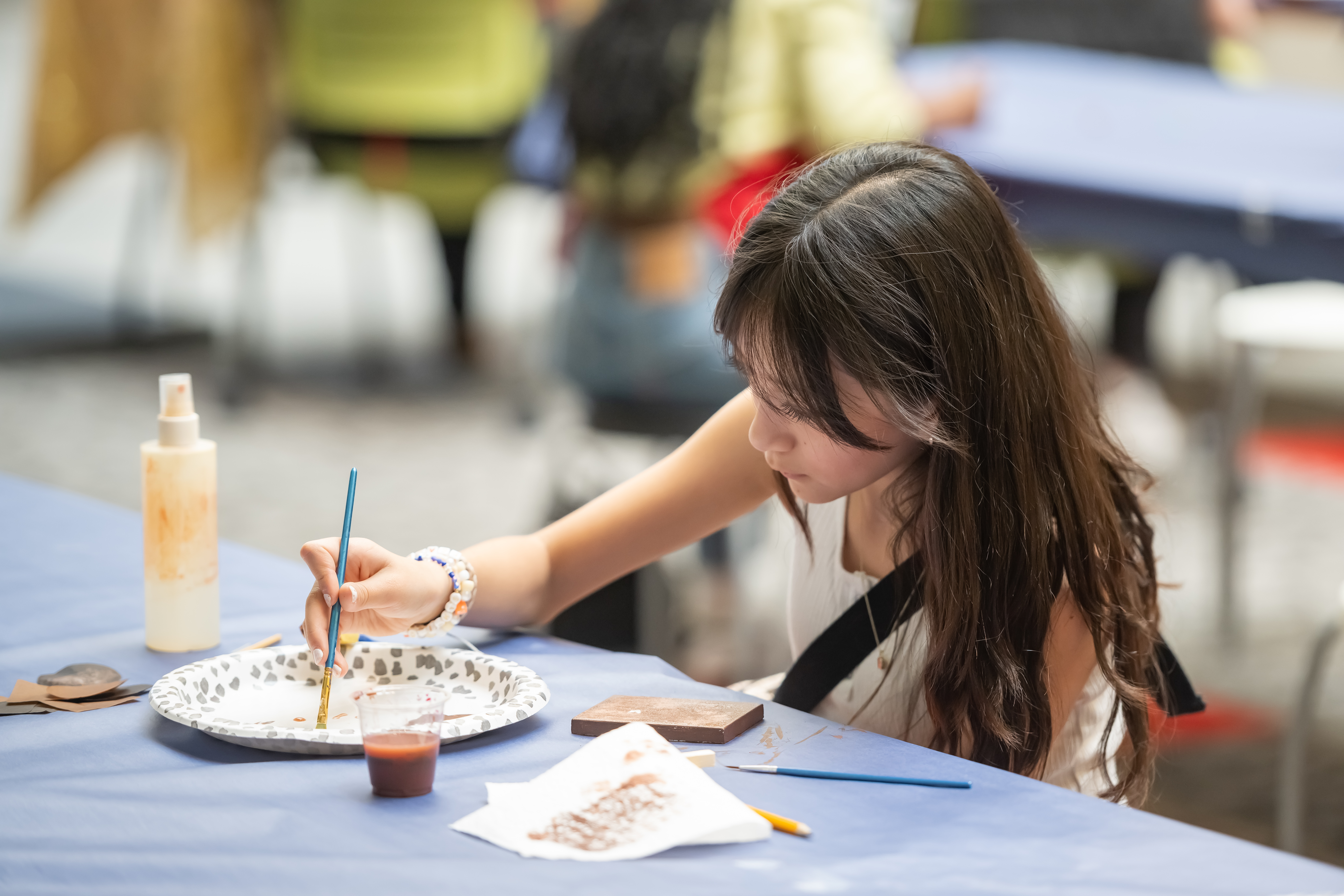 Young light skinned participant with long hair mixing watercolors on a table using a disposable plate to use on a painting she is making.
