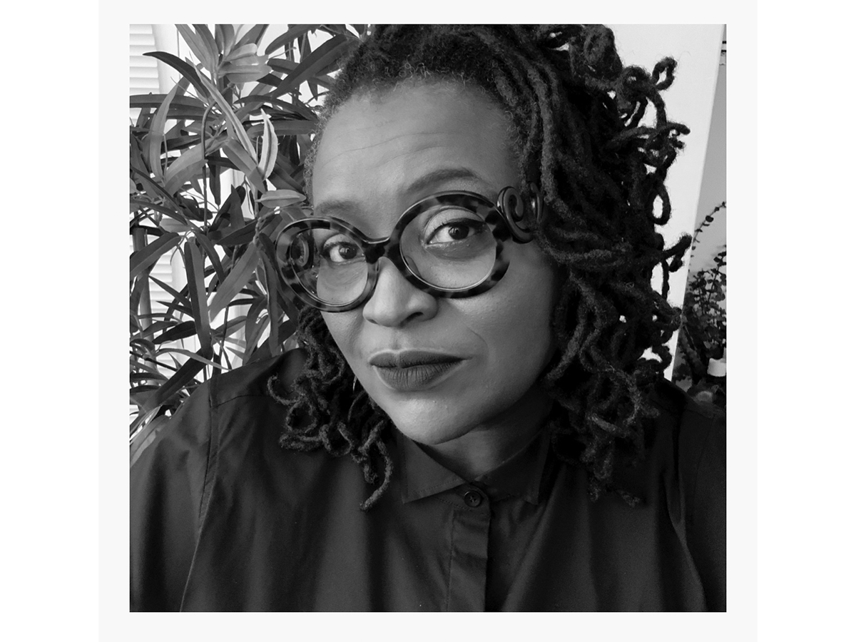 Black and white shoulder/headshot photo of a woman with black rimmed glasses with plants in background