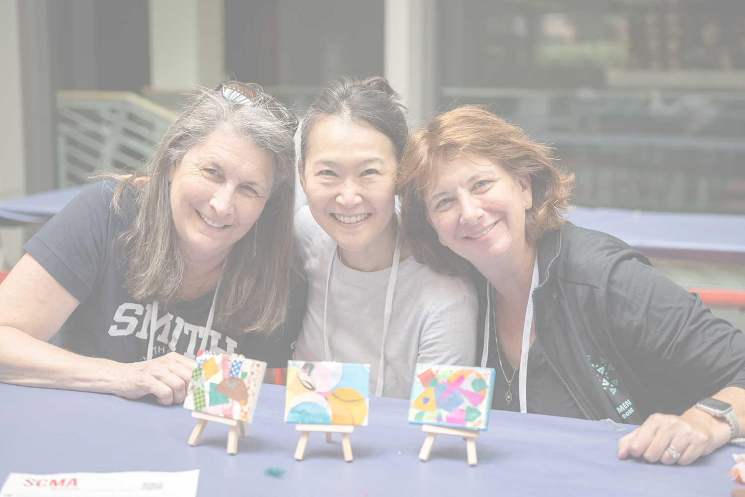 Three medium to light skinned women smiling and posing for the camera while sitting at a table with mini-canvases of art on a mini-easel in front of each person.