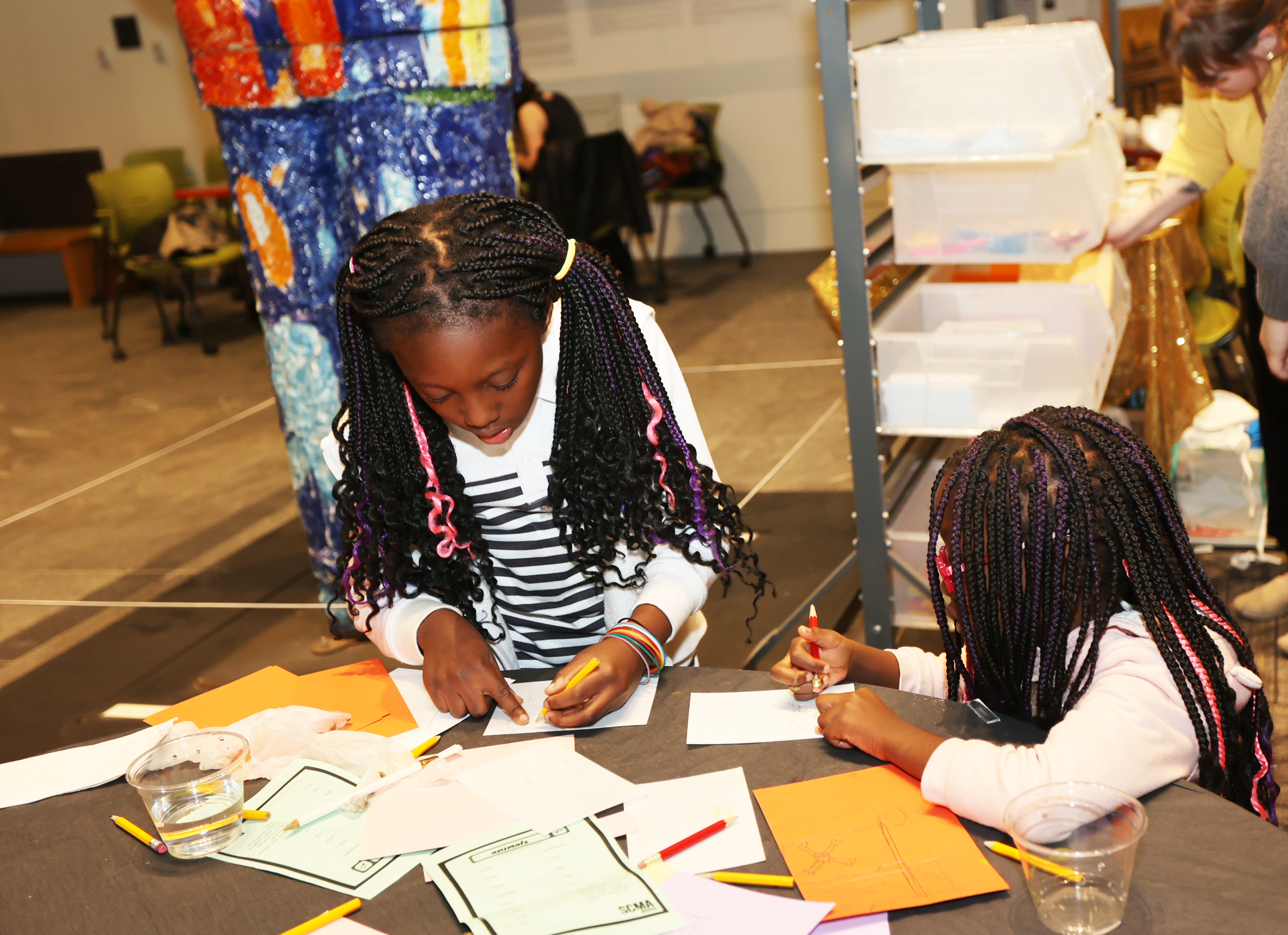 Two young dark skinned girls with braids working at a table drawing together.