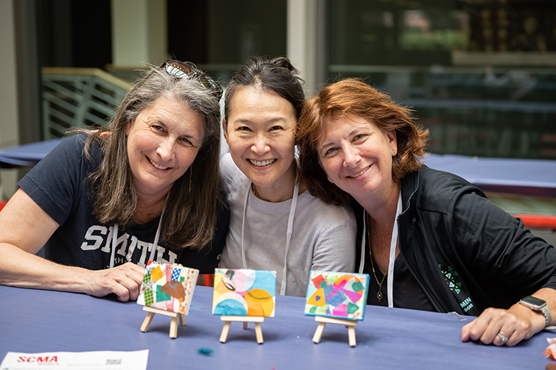 Three women posing at a table with little paintings on them.