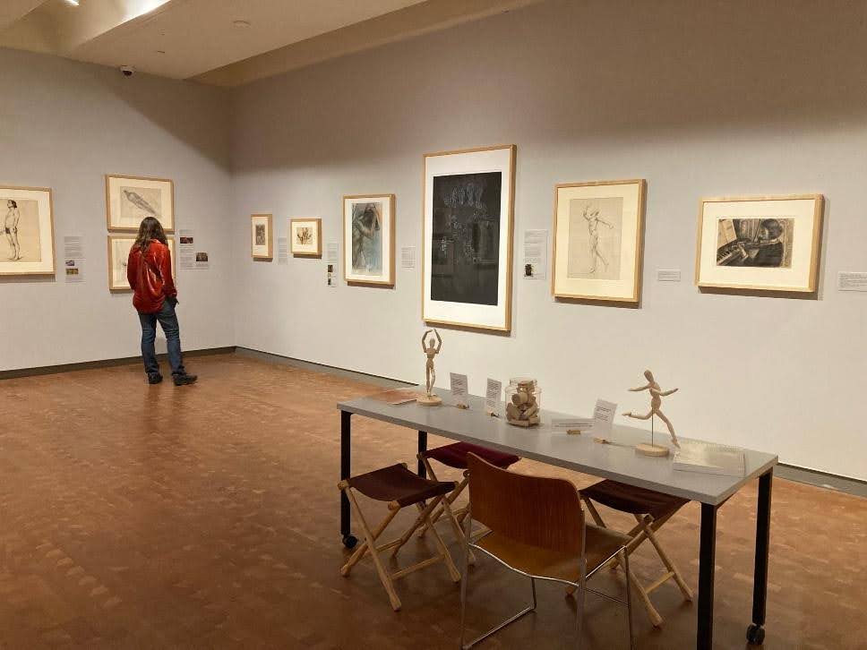 Installation shot of the gallery with person standing on far left looking at art and large table with drawing activity in the foreground.