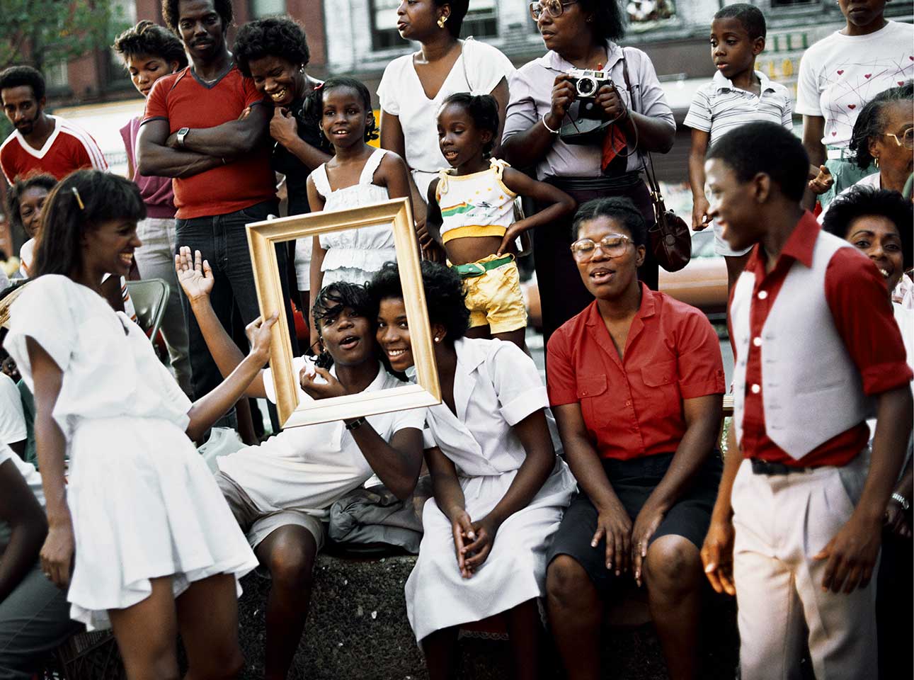 A group of 20 people of color outdoors and dressed in white and red, laughing as a person holds a gold frame in front of the faces of two young kids