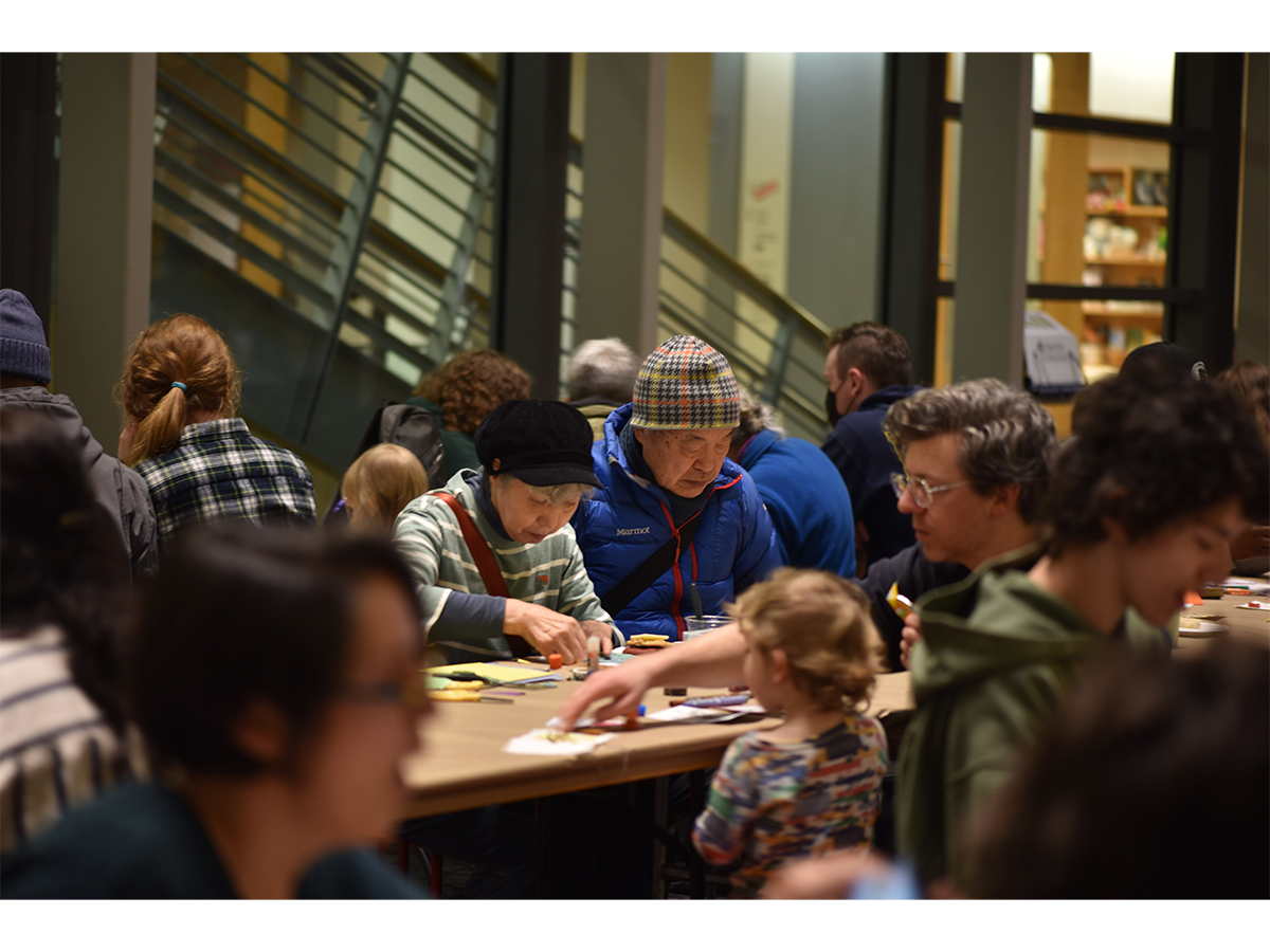 fourteen people dressed in winter clothes sitting at tables making art with paper, scissors and glue