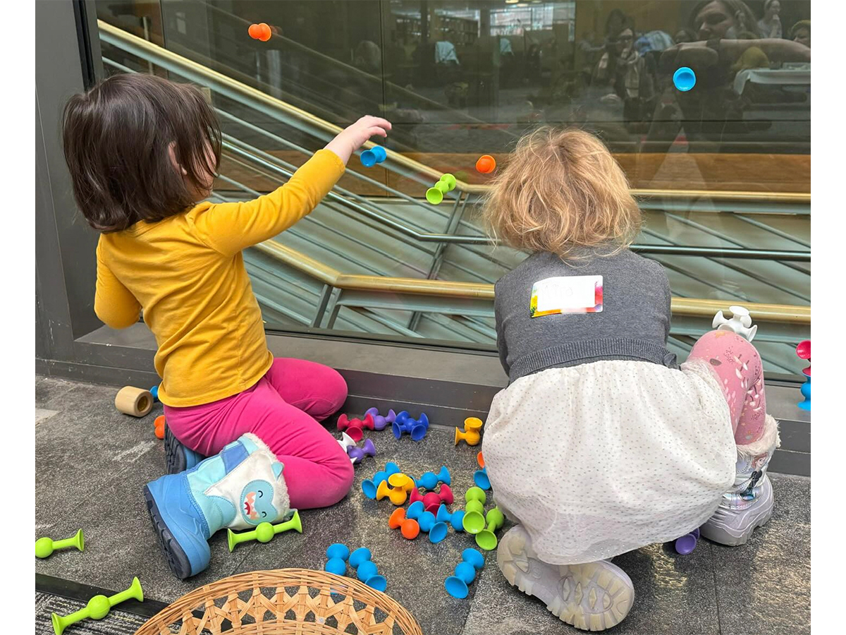Two toddlers playing with sensory toys on a glass wall.