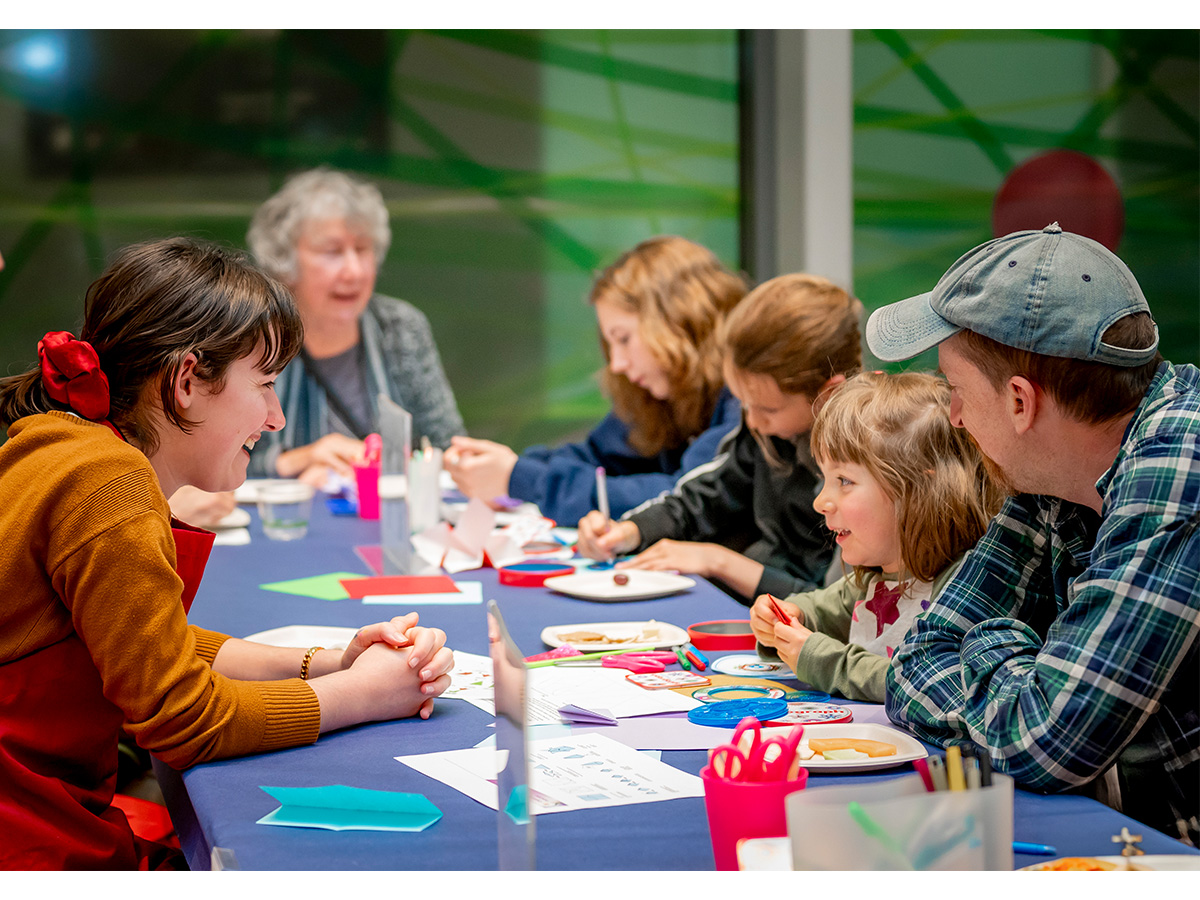 Six people sitting at a table making art, of multiple generations