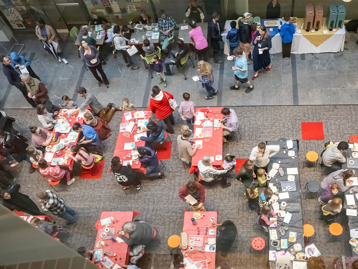 Birds Eye view of a room full of a crowd of people of all ages at tables making art and eating refreshements