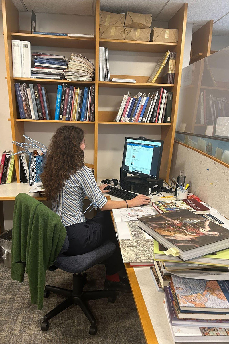 View of Lucy from behind sitting at a desk, typing on a computer and surrounded by books.