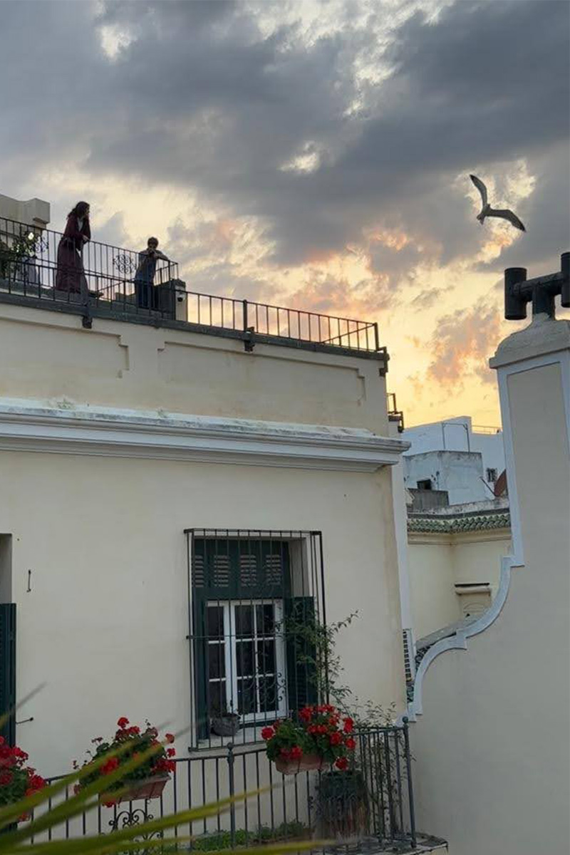 View of a museum facade with two people visible at the top looking out from a rooftop terrace.