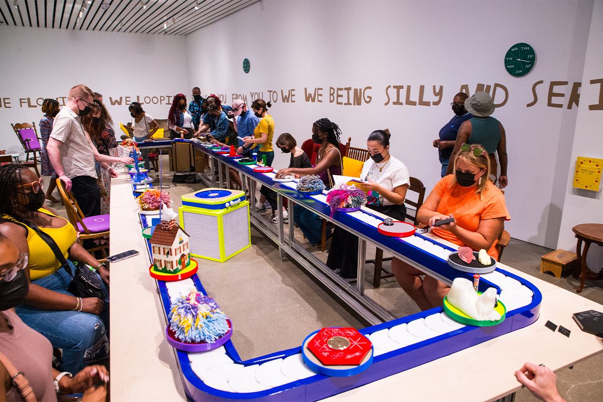 People sit around a conveyor belt interacting with art as it moves around.