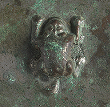 Close-up of a small sitting frog that protrudes from the center of the back of a bronze mirror.
