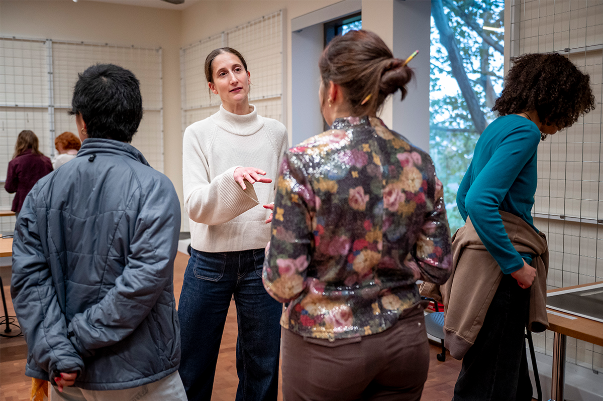 A student in a blue jacket and a student in a pink floral shirt, with their backs to the camera, talk to a person in a white turtleneck. A student on the right looks down at a table just out of view.