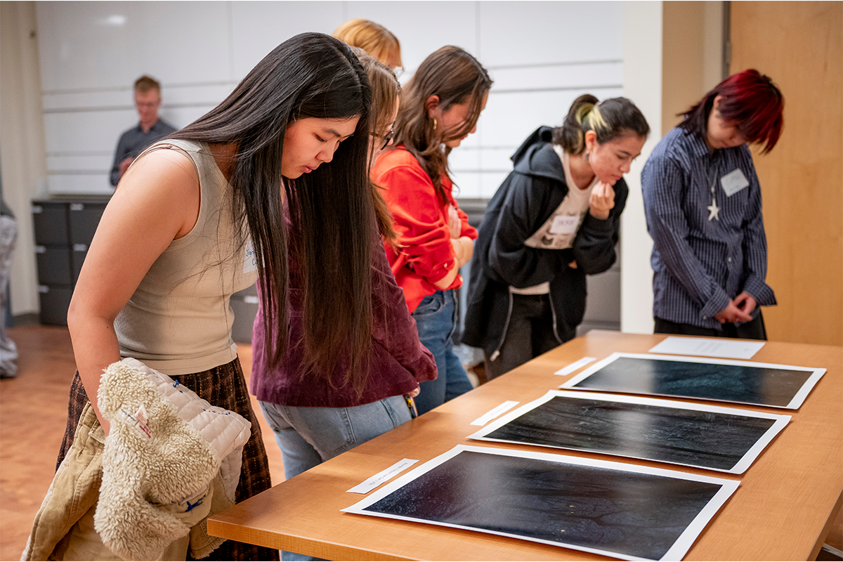 Five students lean over one side of a wooden table and look down at three big, dark photos that lie on top of the table.