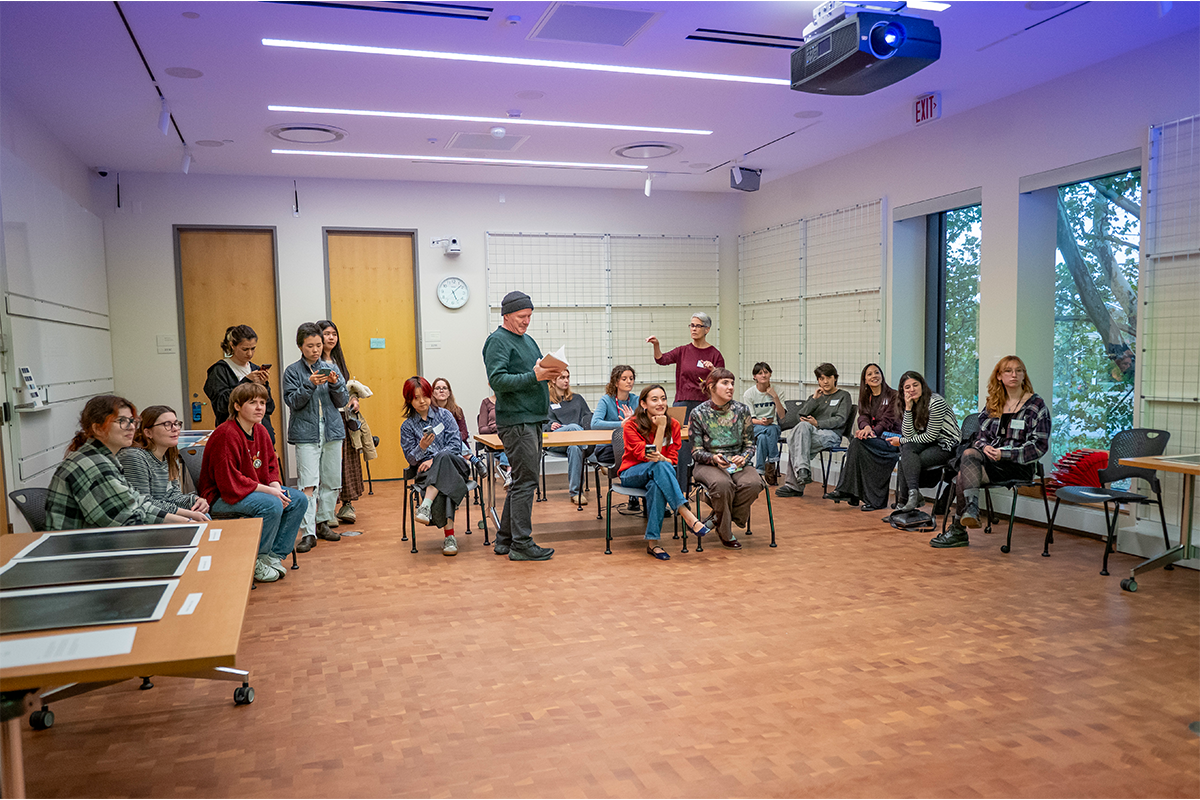 About 18 students sit and stand near the back wall of a big room with their attention directed towards the front of the room. Three printed photographs lie on a table in the lower left corner.