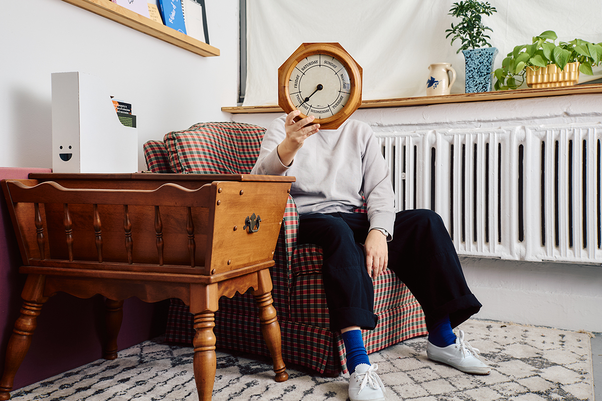 Person in room seated while holding a day of the week clock over their face. There is a table to the left