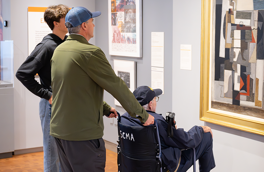 A young person, standing with older person holding the arms of a wheelchair where an elderly person sits, all in front of paintings on a wall in a gallery an elde