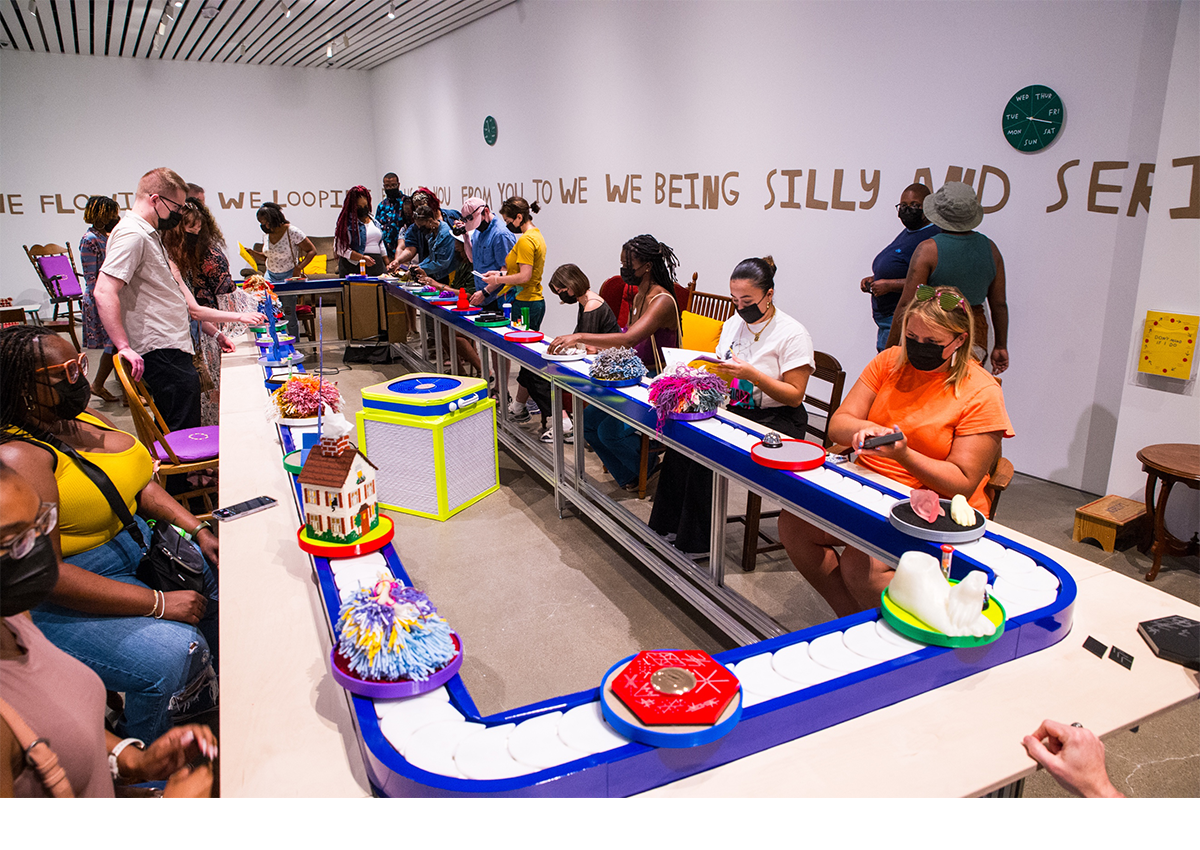Picture of people interacting with an art installation that features text on the walls, and different artworks on discs rotating on a large blue and white conveyor belt. 