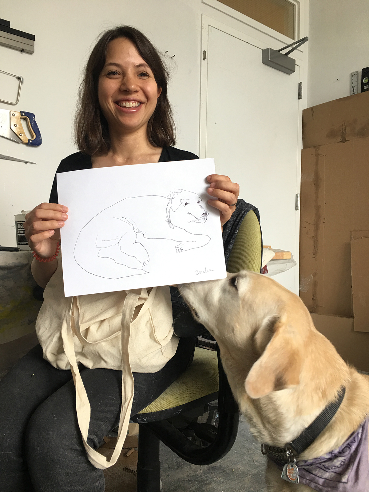 A portrait of Emilie, a white woman with dark brown shoulder-length hair, sits in a green office chair with a tote bag on her lap. She is smiling and holding a sketch of her dog, London.
