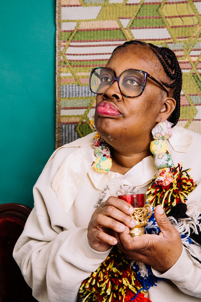 The artist, a Black woman with braids and multicolored glasses, is holding what looks to be a small cup or candle holder.