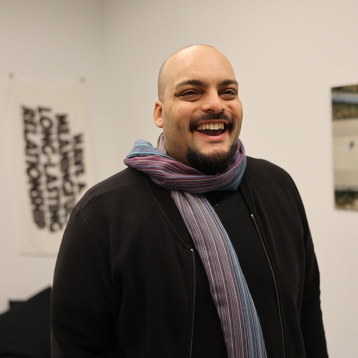 Jeff Kasper smiles warmly in a gallery space. He has a shaved head, a neatly groomed goatee, and is wearing a dark zip-up jacket over a black shirt. 