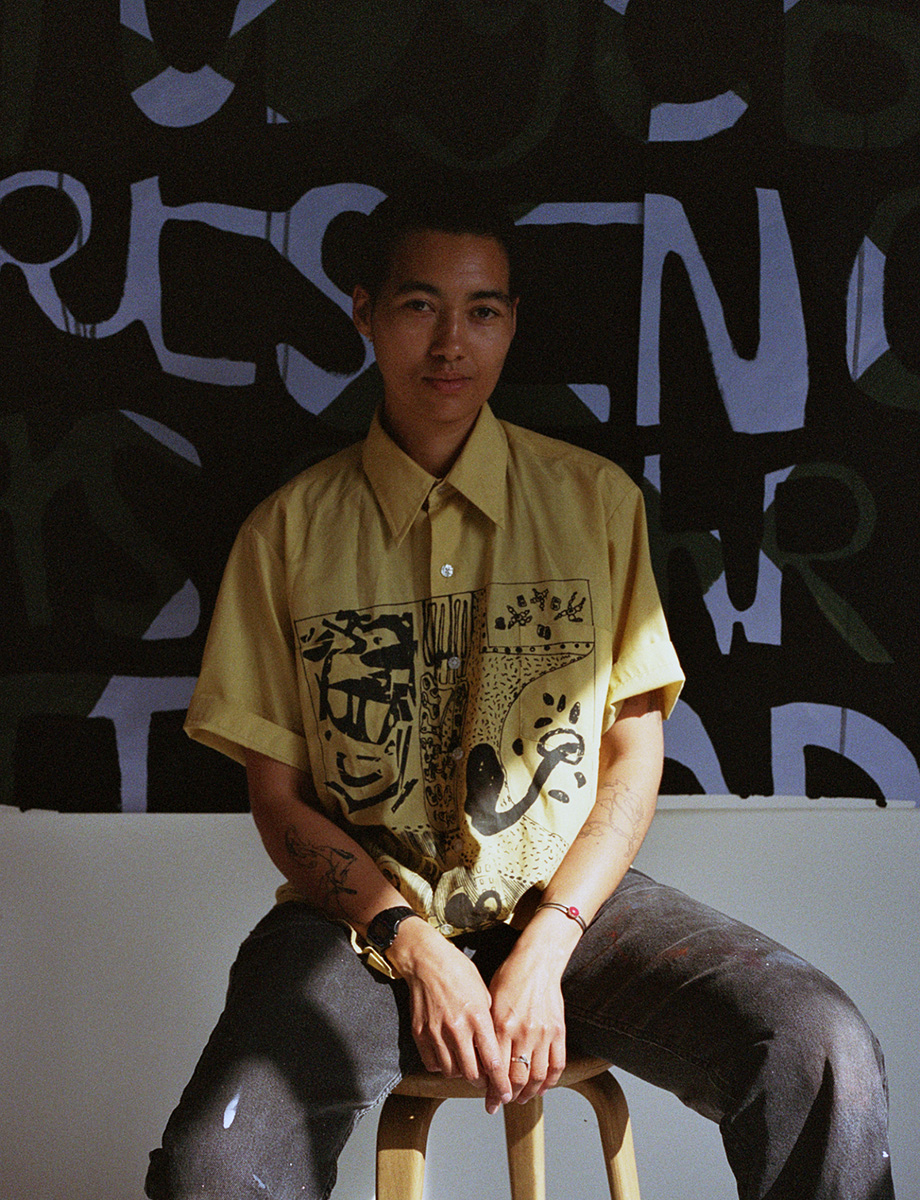 Lukaza Branfman-Verissimo stares directly at the camera with a slight smile. They are sitting in a dimly lit room on a stool with their artwork behind them. Sunlight comes in and shines on their arms. They are wearing a yellow short sleeve button up with a black print on the front.